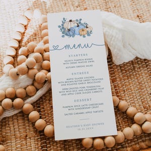 May include: A white rectangular menu card featuring a blue pumpkin illustration at the top and the text: MENU, STARTERS, VELVETY PUMPKIN BISQUE, AUTUMN GREENS SALAD, ENTREES, MAPLE GLAZED CHICKEN, HERB-CRUSTED PORK TENDERLOIN, DESSERT, HEATHER'S BABY SHOWER 10.21.20XX.