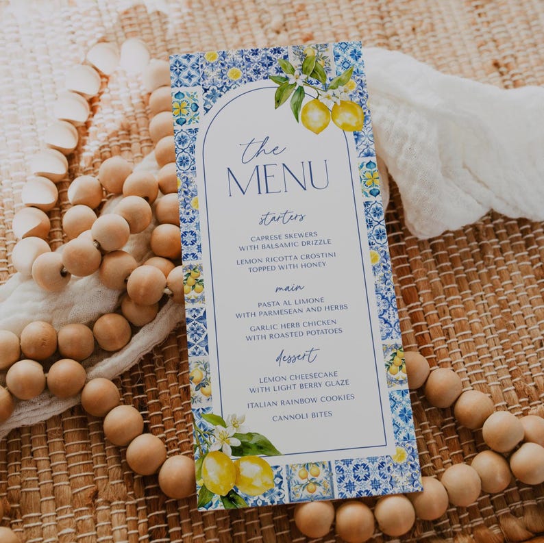 Puede incluir: Una tarjeta de men&uacute; con un dise&ntilde;o de azulejos azules y blancos, con limones y detalles florales. El men&uacute; enumera entrantes, platos principales y postres. El texto del men&uacute; incluye platos como brochetas Caprese y tarta de queso de lim&oacute;n.