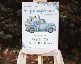 May include: A sign on an easel with the text "Our Sweet Little Pumpkin is Turning One!" and "Welcome to Jayden's 1st Birthday." The sign features a blue truck carrying pumpkins in shades of blue and orange, with floral accents.