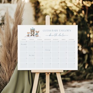 May include: A light blue baby shower calendar with the text "GUESS BABY TAYLOR'S birth date." The calendar features illustrations of a fox, bear, and deer. The calendar is displayed on a wooden easel.