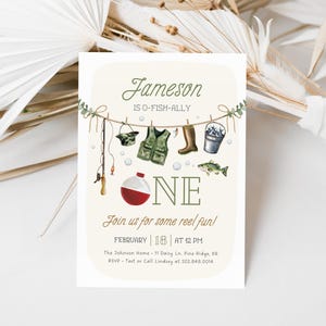 May include: A light beige invitation card with a fishing theme. The card says "Jameson is O-Fish-Ally One" with a fishing rod, a life vest, boots, a bucket of fish, and a fish hanging on a clothesline. The text also says "Join us for some reel fun!" with the date and time of the event, the location, and contact information.