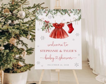 Pode incluir: Uma placa de boas-vindas para chá de bebê em um cavalete. A placa apresenta um vestido vermelho e uma meia pendurados em um varal, com flocos de neve e vegetação. O texto diz "welcome to STEPHANIE & TYLER'S baby shower" e a data.