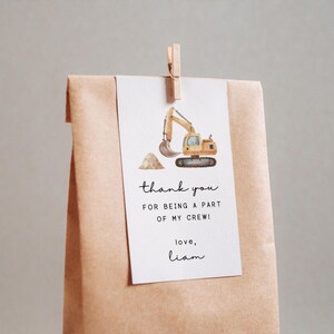 May include: A gift tag featuring a yellow excavator illustration, attached to a brown paper bag with a wooden clip. The tag says "thank you FOR BEING A PART OF MY CREW! love, Liam."