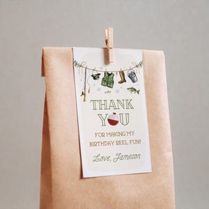 May include: A white thank you card with a fishing theme. The card has a red and white fishing bobber, a fishing rod, a life vest, boots, and a bucket with fishing lures. The text on the card reads "Thank You for making my birthday reel fun! Love, Jameson".