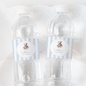 以下が含まれることがあります： 白いキャップが付いた透明なプラスチック製のウォーターボトルが2本。各ボトルには、青いストライプ、テディベアのイラスト、そして「Mila's BABY SHOWER」というテキストが書かれたラベルが付いています。ボトルは白い装飾的な表面に置かれています。