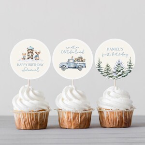 Op de afbeelding: Drie cupcakes met witte frosting en decoratieve toppers. De toppers tonen aquarel illustraties van bosdieren, een blauwe truck en winterbomen. Tekst op de toppers luidt "Happy Birthday Daniel", "Winter ONEdarland" en "Daniel's first birthday".
