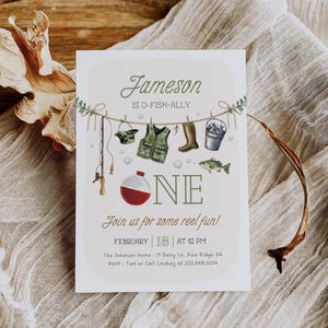 May include: A light blue and green illustrated invitation for a first birthday party. The invitation features a fishing theme with a fishing rod, a life vest, boots, a bucket, and a fish. The text on the invitation reads "Jameson is O-Fish-Ally One. Join us for some reel fun! February 18 at 12 PM. The Johnson Home 11 Daisy Ln. Pine Ridge, PA. RSVP - Text or Call Lindsey at 202.848.0014."