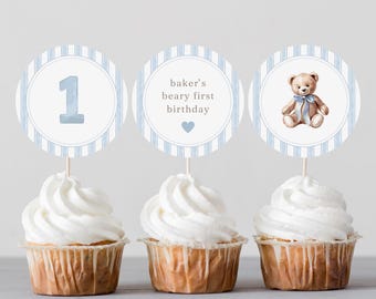 May include: Three cupcakes with white frosting and decorative toppers. The toppers feature a blue number 1, the text "baker's beary first birthday" with a heart, and a teddy bear with a blue bow. The toppers have a blue and white striped border.