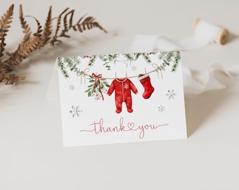 Könnte beinhalten: Gefaltete Grußkarte mit einer Aquarellillustration eines roten Baby-Stramplers und Strumpfes, die an einer Wäscheleine hängen, mit Grün und Schneeflocken. Die Worte "Danke" sind in roter Schrift geschrieben.