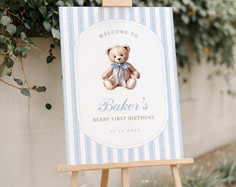 May include: A welcome sign for a first birthday party. The sign features a watercolor illustration of a teddy bear with a blue bow tie. The sign has a blue and white striped background and reads "Welcome to Baker's Beary First Birthday".