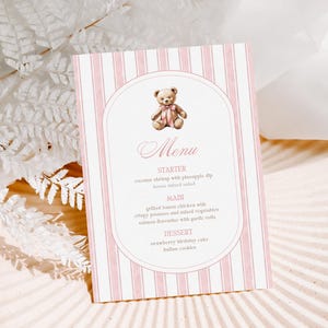 May include: A menu card with pink and white stripes, adorned with a teddy bear illustration. The menu lists starters, main courses, and desserts. The card is presented on a light surface with decorative details.