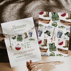 May include: A white invitation with a watercolor illustration of fishing gear hanging on a clothesline. The text reads "Jameson is O-Fish-Ally One. Join us for some reel fun! February 18 at 12 PM. The Johnson Home 11 Daisy Ln. Pine Ridge, PA. RSVP - Text or Call Lindsey at 202.848.0014."