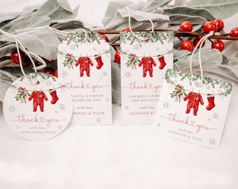 May include: A collection of white gift tags with a Christmas theme. Each tag features a red onesie and stocking design, with the words "thank you" in red script. The tags are tied with string and decorated with greenery and snowflakes.