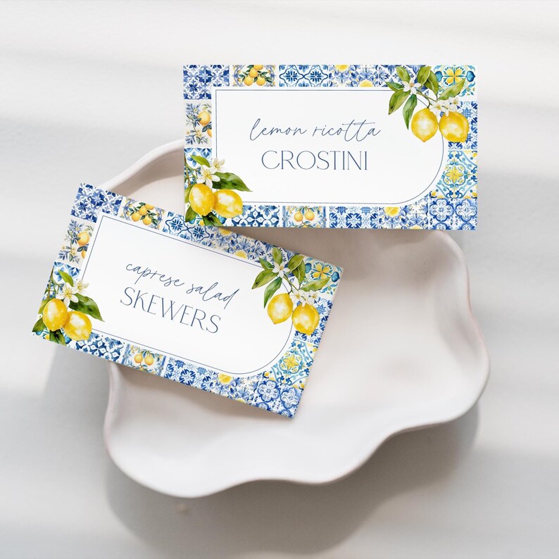 Puede incluir: Dos tarjetas rectangulares con un patr&oacute;n de azulejos azules y blancos e ilustraciones de limones. Una tarjeta dice "lemon ricotta CROSTINI" y la otra dice "caprese salad SKEWERS". Las tarjetas est&aacute;n sobre un plato blanco con borde festoneado.