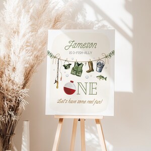 May include: A white sign with a green and brown fishing theme. The sign says "Jameson is O-Fish-Ally One. Let's have some reel fun!"