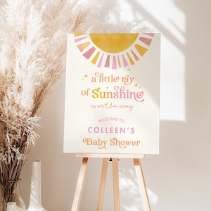 May include: A white sign with a yellow and pink sun graphic. The text reads "a little ray of Sunshine is on the way. Welcome to Colleen's Baby Shower."