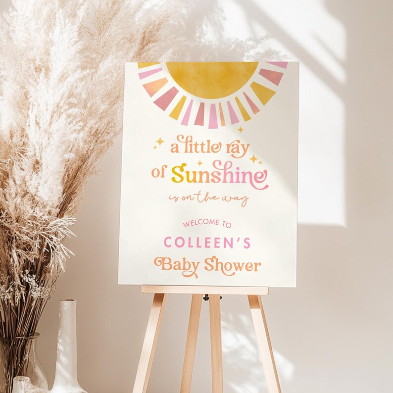 May include: A white sign with a yellow and pink sun graphic. The text reads "a little ray of Sunshine is on the way. Welcome to Colleen's Baby Shower."