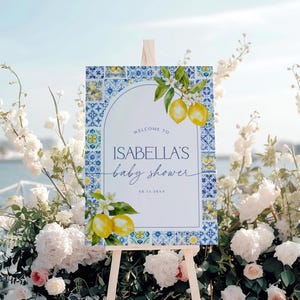 Puede incluir: Un letrero de bienvenida para baby shower sobre un caballete de madera. El letrero presenta un dise&ntilde;o de azulejos azules y blancos con limones y el texto "Welcome to Isabella's baby shower". El letrero est&aacute; rodeado de flores blancas y vegetaci&oacute;n.