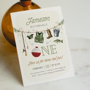 May include: A fishing-themed first birthday invitation for a boy named Jameson. The invitation features a green, white, and red fishing bobber, a fishing rod, a fishing vest, and a bucket of fish. The text reads "Jameson is O-Fish-Ally One. Join us for some reel fun! February 18 at 12 PM. The Johnson Home 11 Daisy Ln. Pine Ridge, PA. RSVP Text or Call Lindsey at 202.848.0014."
