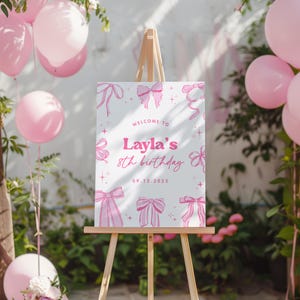 May include: A wooden easel displays a white sign with pink bows and the text "Welcome to Layla's 8th birthday 09.12.2025." Pink balloons and greenery surround the sign, creating a festive atmosphere.