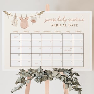 Peut inclure: Un calendrier blanc avec une grille brune et le texte "guess baby carter's arrival date". Le calendrier est décoré d'une ligne de vêtements avec une barboteuse rose pour bébé, un bouquet de fleurs et un chapeau de bébé.