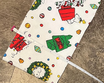 May include: White fabric with a Christmas-themed pattern featuring Snoopy, wrapped gifts, Christmas trees, and ornaments. A measuring tape shows the fabric is approximately 6 inches wide.