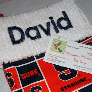 May include: A white stocking with the name "David" embroidered on it. The stocking is decorated with a red and blue pattern featuring the word "CUSE" and the word "SYRACUSE". A small tag is attached to the stocking with the text "Holly's Baby Holly Jolly Holidays Boutique Christmas Stocking holly jaquith creator PERSONALIZATION AVAILABLE 100% of fabrics to choose from".