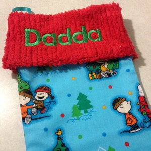 May include: A Christmas stocking with a red cuff embroidered with the word "Dadda" in green. The body of the stocking is light blue and features a pattern of cartoon characters, Christmas trees, and colorful dots.