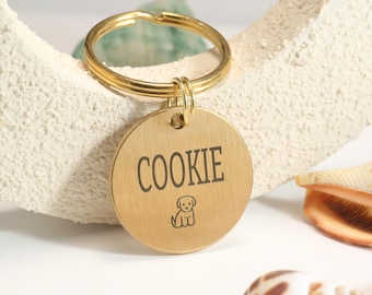 May include: A gold-colored, round pet tag with the name "COOKIE" in black lettering and a small dog illustration. The tag is attached to a gold-colored split ring. The background includes a white, textured surface and seashells.