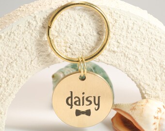 May include: A gold pet tag with the name "daisy" in a stylish font and a small bow tie graphic. The tag is attached to a gold keyring. The background includes a white, textured object, a green stone, and a seashell.