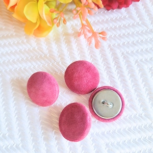 May include: Four round, magenta fabric-covered buttons. One button is open, showing the metal back and shank. The buttons are on a white textured surface, with colourful flowers in the background.
