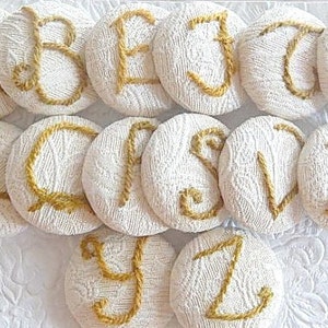 May include: A set of 12 white fabric buttons with gold embroidered letters spelling out the alphabet from A to L. The buttons are arranged in a circle on a white surface.