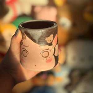 May include: A ceramic mug with a hand-painted design. The mug features a cartoon face with large eyes, rosy cheeks, and a small bear illustration. The top of the mug is dark brown, and the face is light pink.