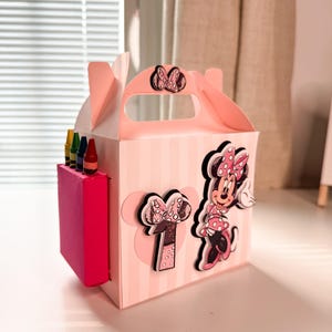 Puede incluir: Caja de regalo rosa y blanca con un personaje de Minnie Mouse y un número uno en la parte delantera. La caja tiene un asa y está llena de lápices de colores.