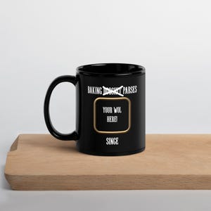 May include: Black ceramic mug with a white handle. The mug features the text "BAKING DOUGHNUT PARSES" above a gold-bordered rectangle with "YOUR WOL HERE!" and "SINCE" in white text. The mug sits on a wooden surface.