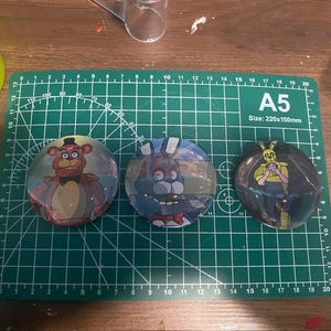 May include: Three round, clear, domed buttons featuring characters from the video game series Five Nights at Freddy's. The buttons are set on a green cutting mat with grid lines. The characters are Freddy Fazbear, Bonnie, and Springtrap.