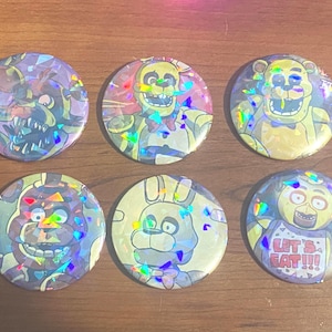 May include: Six circular pin-back buttons featuring holographic foil overlays and illustrations of various animatronic characters. One button displays a yellow chicken character with a bib that has the text LET'S EAT!!! printed in red letters.