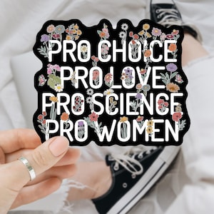 Può includere: Un adesivo nero con testo bianco che recita "PRO CHOICE, PRO LOVE, PRO SCIENCE, PRO WOMEN". L'adesivo è decorato con fiori e farfalle colorate. L'adesivo è tenuto in mano con un anello d'argento.