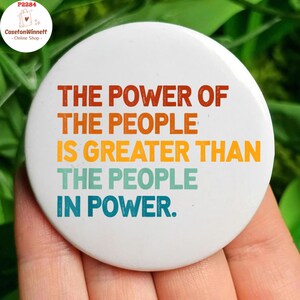 May include: A white button with a colourful quote that reads "THE POWER OF THE PEOPLE IS GREATER THAN THE PEOPLE IN POWER."