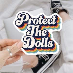 May include: A sticker with the text "Protect The Dolls" in a retro font, outlined in white and layered over a rainbow design. The sticker is held by a person, with a laptop and black sneakers in the background.