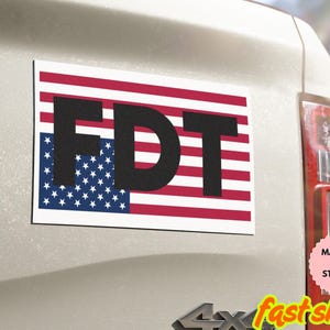 May include: Rectangular magnet or sticker with the letters "FDT" in bold black font over an American flag design. The flag features red and white stripes and a blue field with white stars. The magnet is affixed to a vehicle.