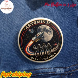 May include: A round pin button featuring a space-themed design. The button displays the text "ARTEMIS II" with a rocket launching towards the moon, astronauts, and the text "BACK TO THE MOON". The button is set against a dark blue background.