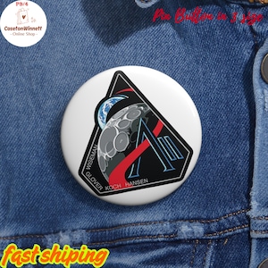 May include: A white pin button with a black, red, and blue graphic design featuring a stylized moon and Earth. The button has the names "Wiseman", "Glover", "Koch", and "Hansen" printed on it. The button is on a denim background.