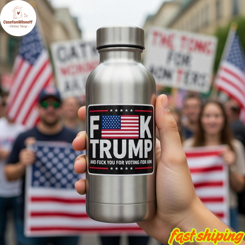 May include: A stainless steel water bottle with a sticker that reads "F**k Trump" with an American flag design. The text "And F**k You For Voting For Him" is below the flag. The bottle is held in front of a crowd of people.