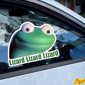 May include: A green lizard sticker is affixed to a car window. The sticker features a cartoon lizard face with large eyes and the text "Lizard Lizard Lizard" in green. The sticker is water-resistant.