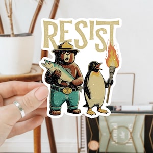 May include: A sticker featuring a cartoon illustration of a bear wearing a ranger hat holding a fish and a penguin holding a torch. The text "RESIST" is above the characters.