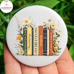 Può includere: Un bottone rotondo bianco con un design di una pila di libri con le parole "Fantasy", "Drama", "Tragedy" sui dorsi. I libri sono circondati da fiori bianchi e gialli.