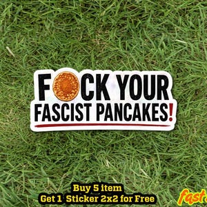 May include: A rectangular sticker featuring the text FOCK YOUR FASCIST PANCAKES! in bold black capital letters. The letter O in FOCK is replaced by a circular image of a golden-brown pancake. A thin horizontal red line is positioned below the text.