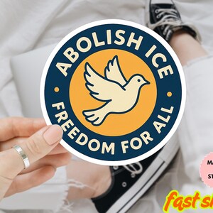 Può includere: Un adesivo rotondo con la scritta "ABOLISH ICE FREEDOM FOR ALL" in blu navy. Una colomba color crema è al centro, su un cerchio giallo. L'adesivo è tenuto da una mano.