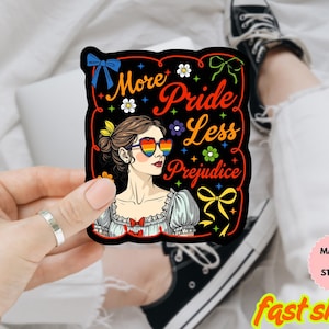 Puede incluir: Un imán o pegatina negra con el texto "More Pride Less Prejudice" y un retrato de una mujer con gafas de sol en forma de corazón arcoíris. El diseño incluye flores, lazos y elementos decorativos.
